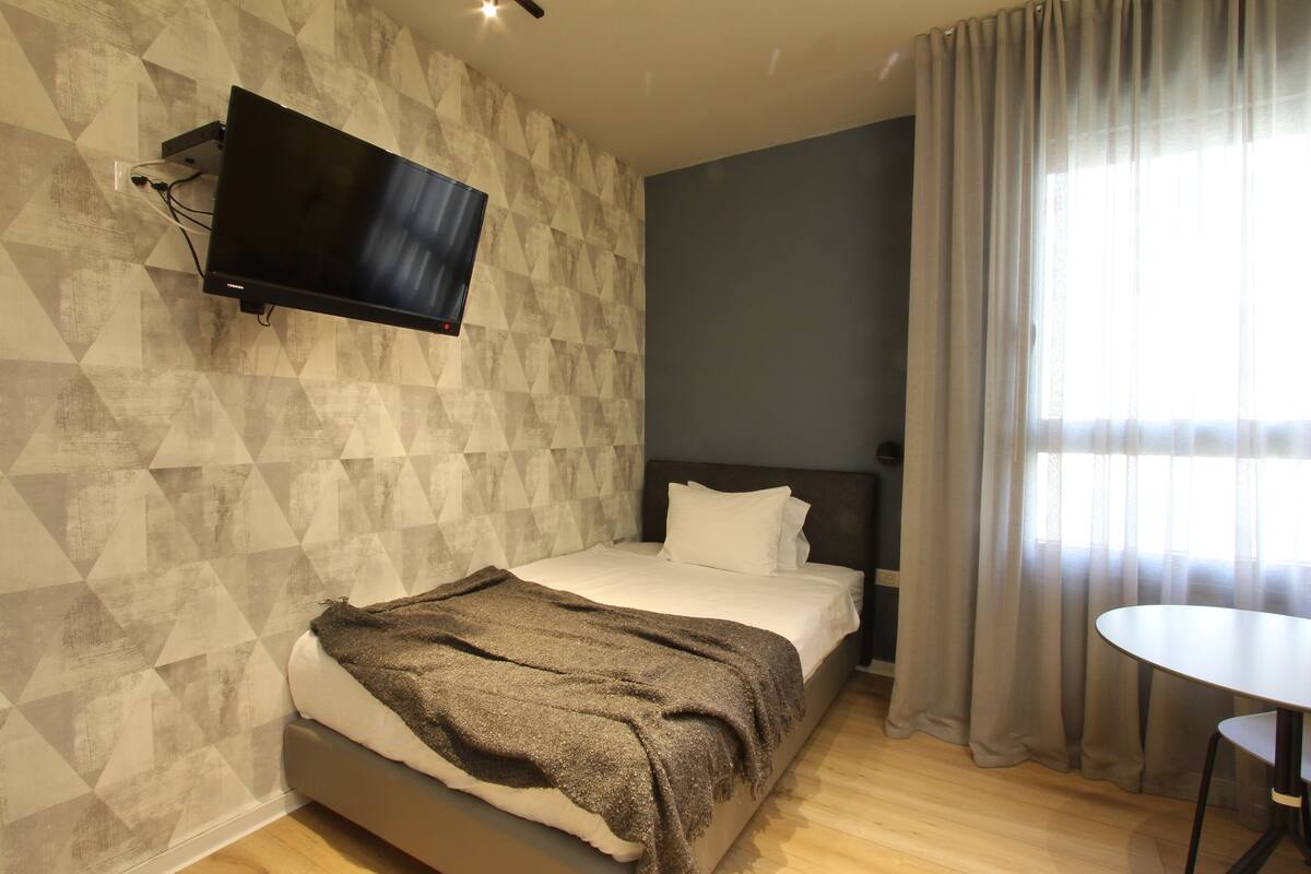 Una habitación de hotel moderna con una cama, un televisor montado en la pared, una mesita y un ventanal con cortinas.
