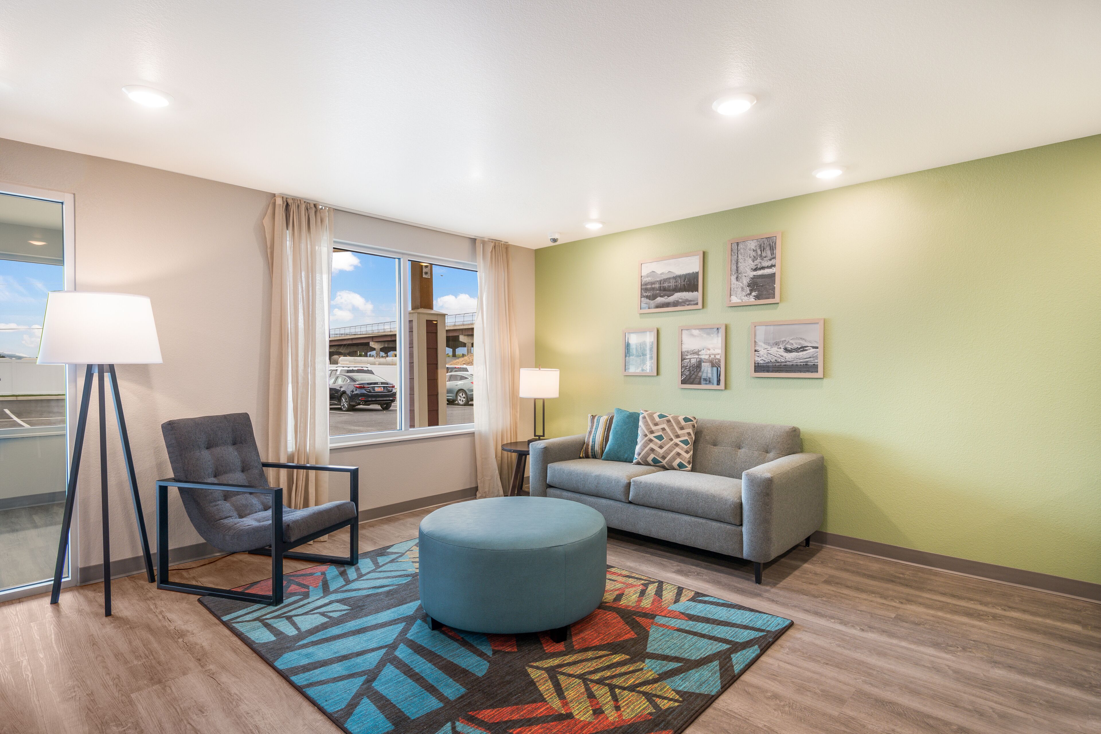 Una sala de estar moderna con sofá, sillón, reposapiés y un amplio ventanal con vistas a un aparcamiento.