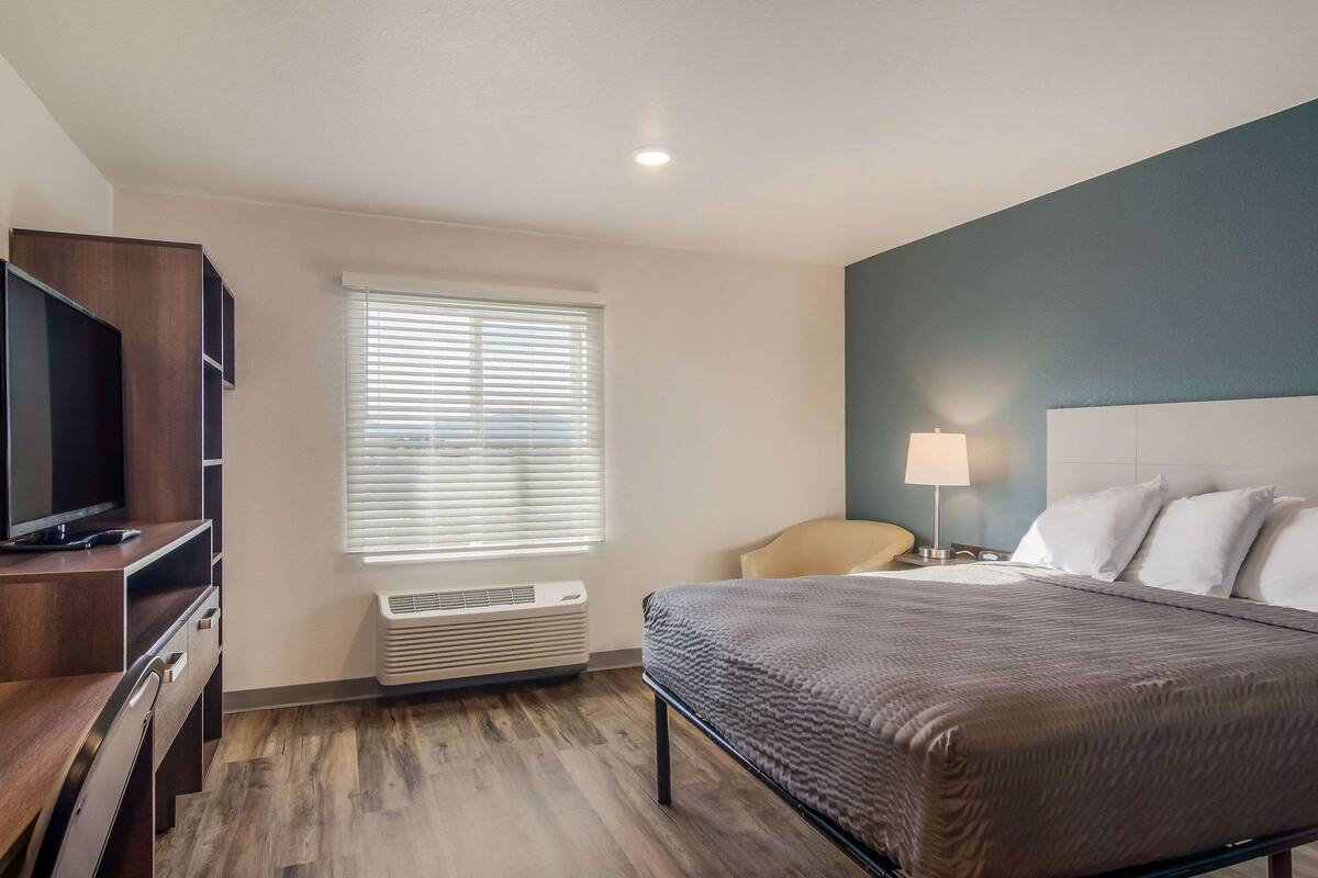 Habitación de hotel con cama, televisor, escritorio, silla y ventana con persianas.