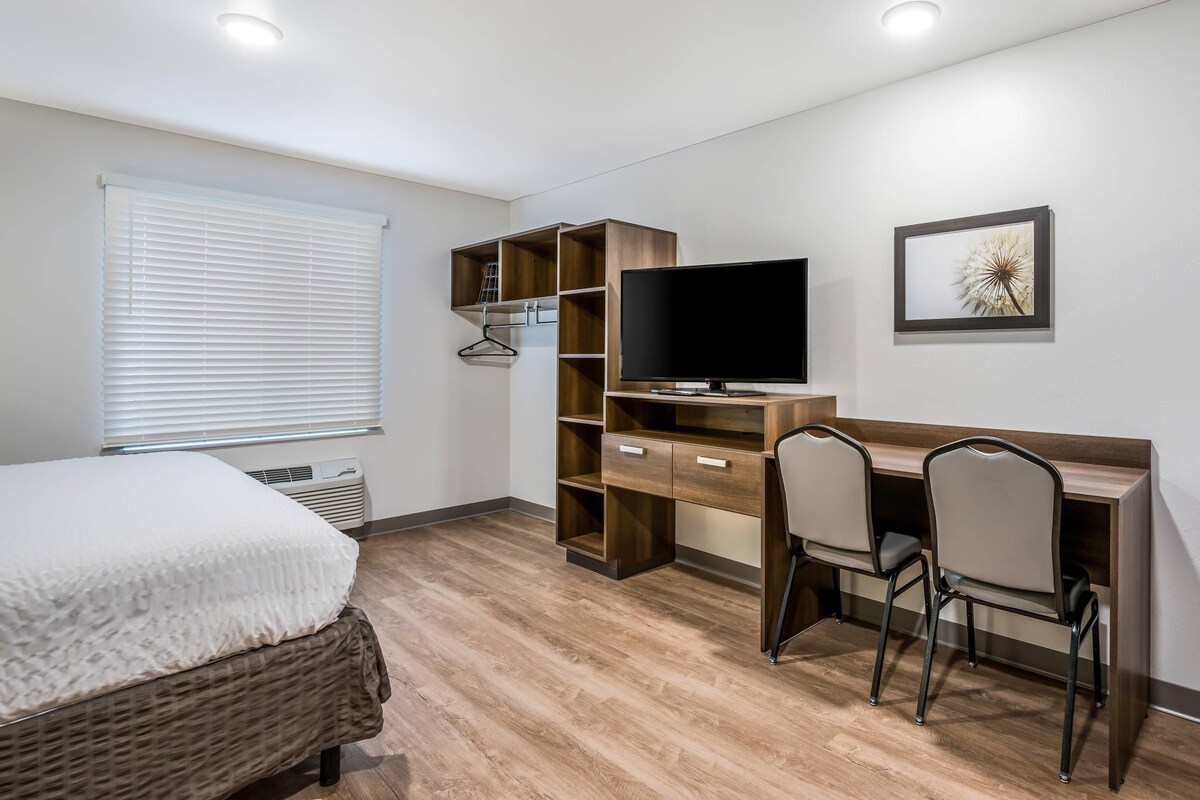 A bedroom with a bed, a desk with a chair, a TV, and a window with blinds.