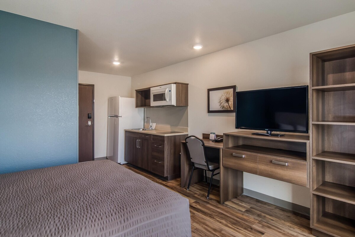 A compact room with a bed, kitchenette, desk, chair, and a TV.