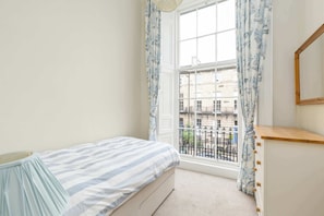 A bedroom with a bed, a window with curtains, a dresser, and a view of buildings outside.