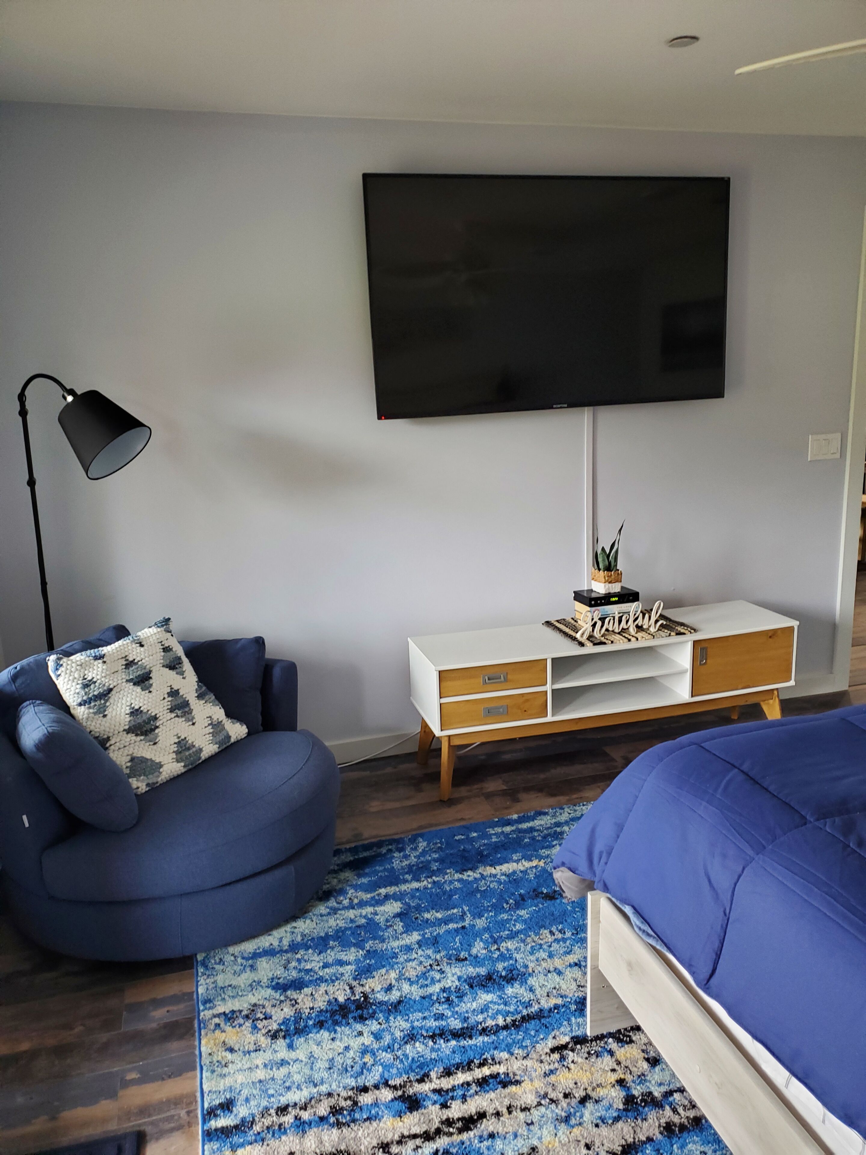 Un dormitorio moderno con una cama, un sillón azul, una mesita de consola blanca, un televisor y una lámpara de pie.