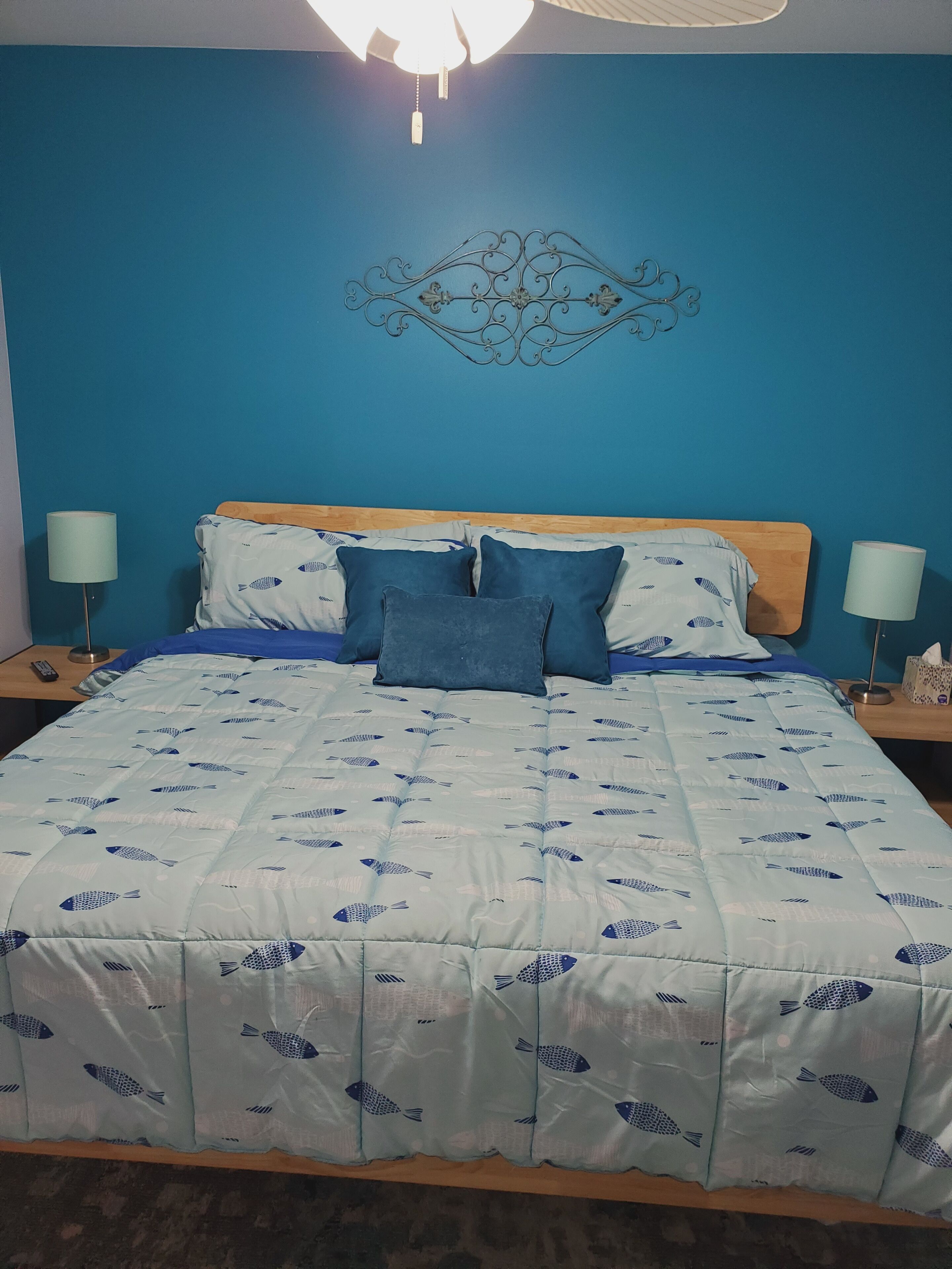 A bed with a blue and white fish-patterned duvet, two pillows, a wooden headboard, and a decorative wall piece above the bed.