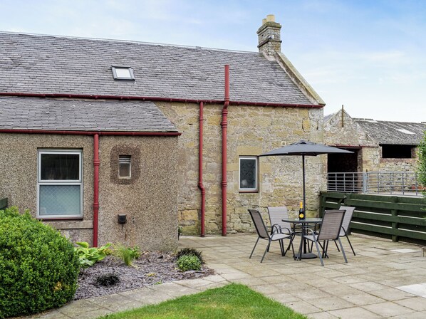 A stone building with a tiled roof, a small window, and a patio with a table and chairs.