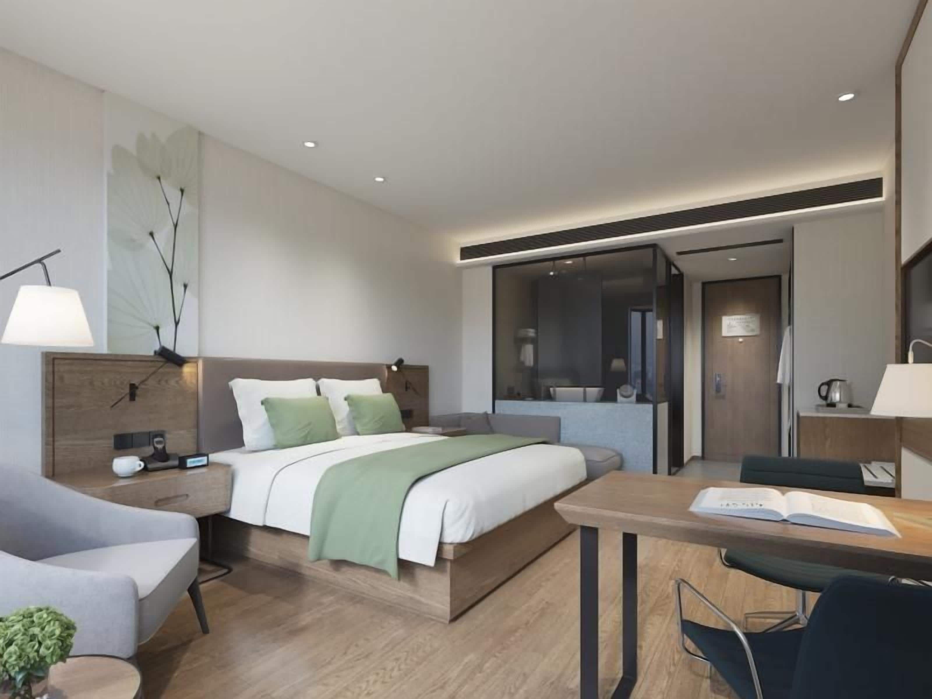 A modern hotel room with a bed, bedside table, lamp, and a desk with a chair.