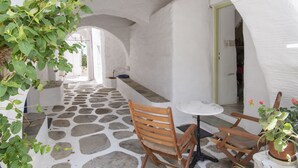 A stone-floored corridor with wooden chairs and a table, white walls, and a doorway leading to another room. - Traditional stonebuilt house (Paros)