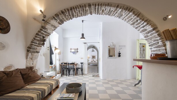 A cozy living room with a checkered floor, a sofa, a table with books, and a stone archway leading to another room. - Traditional stonebuilt house (Paros)
