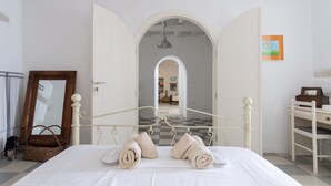 A bedroom with a checkered floor, a bed with rolled towels, a wooden-framed mirror, a wicker basket, and a desk with a chair. - Traditional stonebuilt house (Paros)