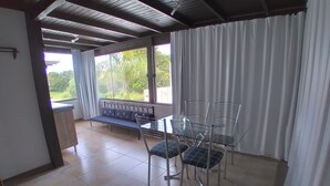 Un patio couvert avec une table et des chaises en verre, un banc avec des coussins et une vue sur la verdure environnante.