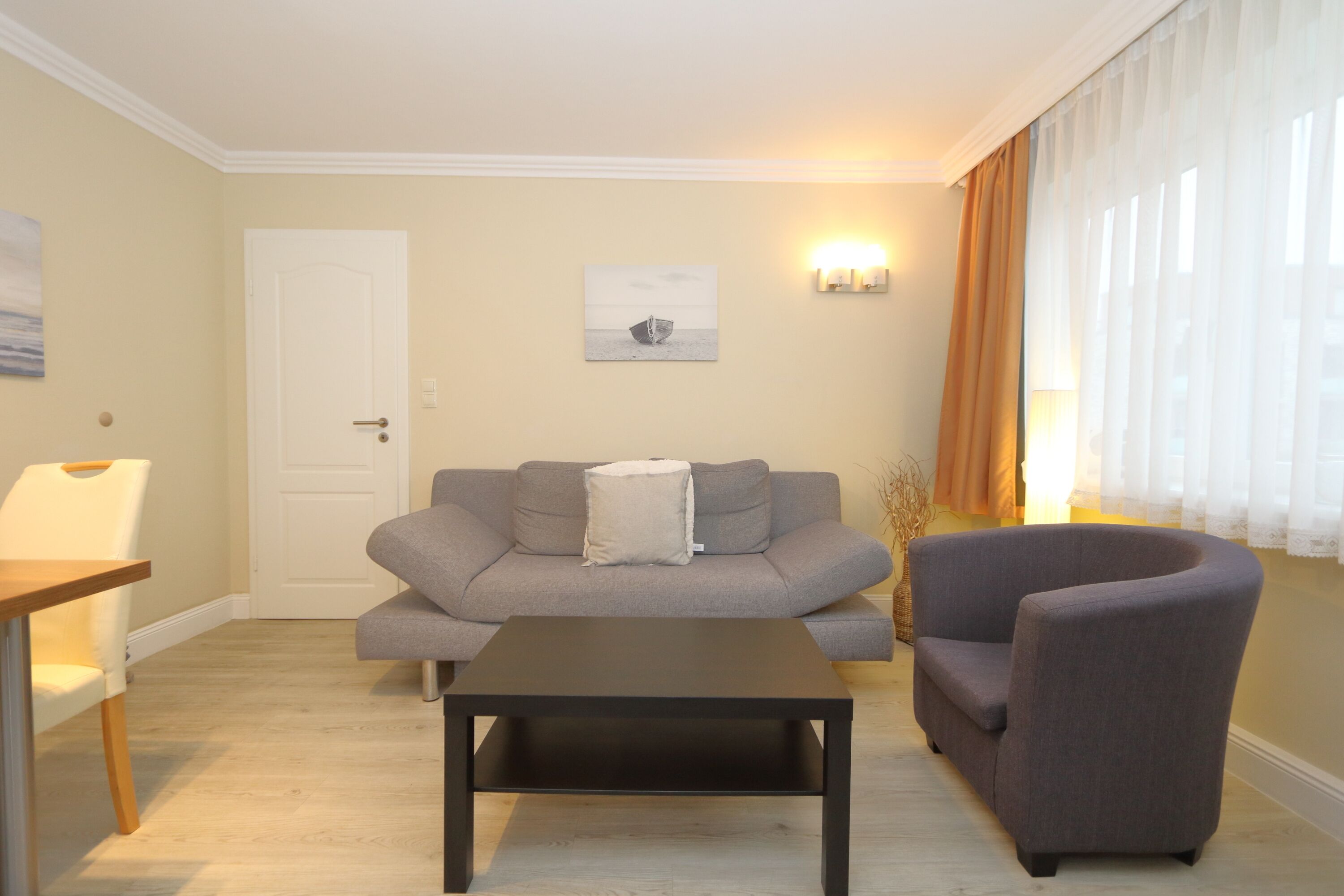 A living room with a grey sofa, a black coffee table, and a grey armchair. There is a white door, a painting on the wall, and a window with curtains.
