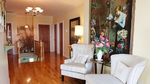 Un salón amplio con suelo de madera, un sillón a rayas, un sillón blanco, una mesita con una lámpara y un cuadro floral grande en la pared.