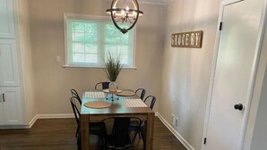 A dining area with a wooden table and chairs, a chandelier, a window with blinds, and a 'GROCERY' sign on the wall. - Convenient 3 Bedroom / 2 Bath house (Gainesville)