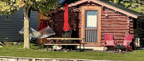 Una cabaña de troncos con un porche, una hamaca, una mesa y dos sillas Adirondack.