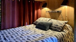Une pièce de cabane en bois comprenant un lit, des rideaux rouges, une étagère avec divers objets et une lampe.