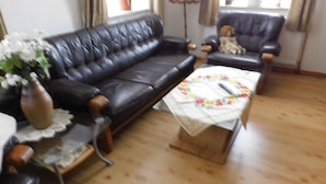 Een woonkamer met een zwarte leren bank, een bijpassende fauteuil, een houten salontafel met een bloemen tafelkleed en een vaas met bloemen.