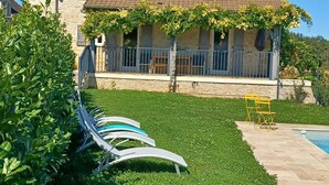 Una casa con techo de tejas, un balcón con plantas trepadoras, una piscina y un césped con sillas de descanso.