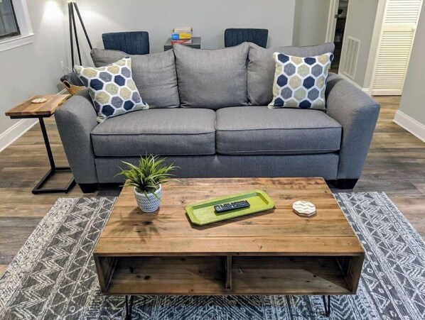 A grey sofa with patterned cushions, a wooden coffee table with a plant and remote, and a patterned rug.