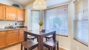 A kitchen with wooden cabinets, a dining table with chairs, a hanging light fixture, and windows with blinds.