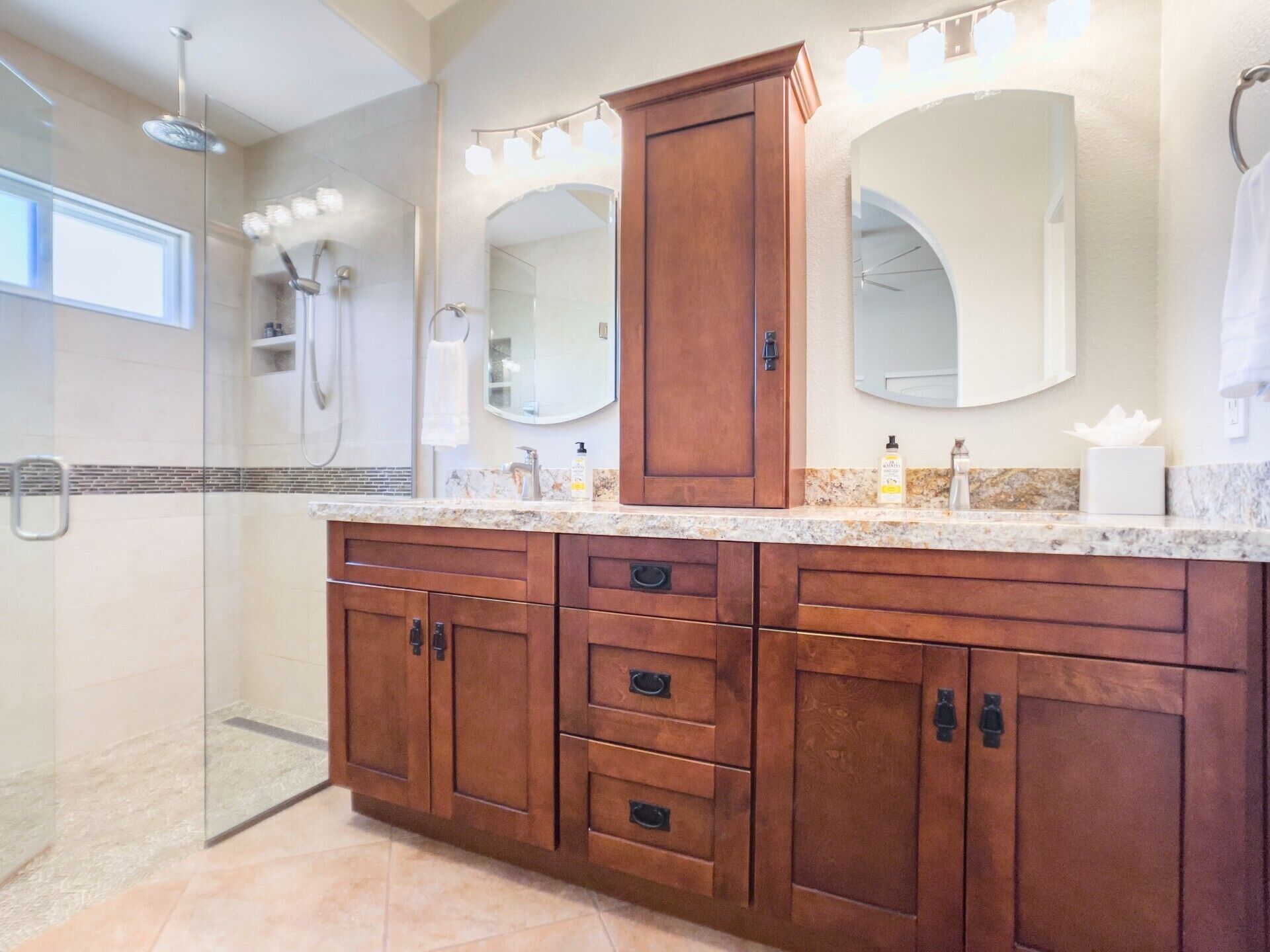 A bathroom with a glass shower enclosure, wooden vanity with multiple drawers, and two large mirrors.