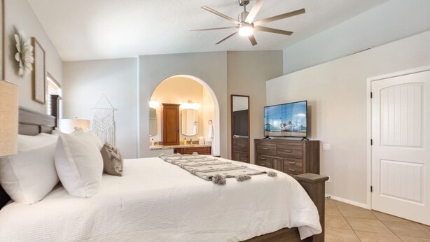 A bedroom with a bed, a ceiling fan, a TV, and a bathroom visible through an archway.