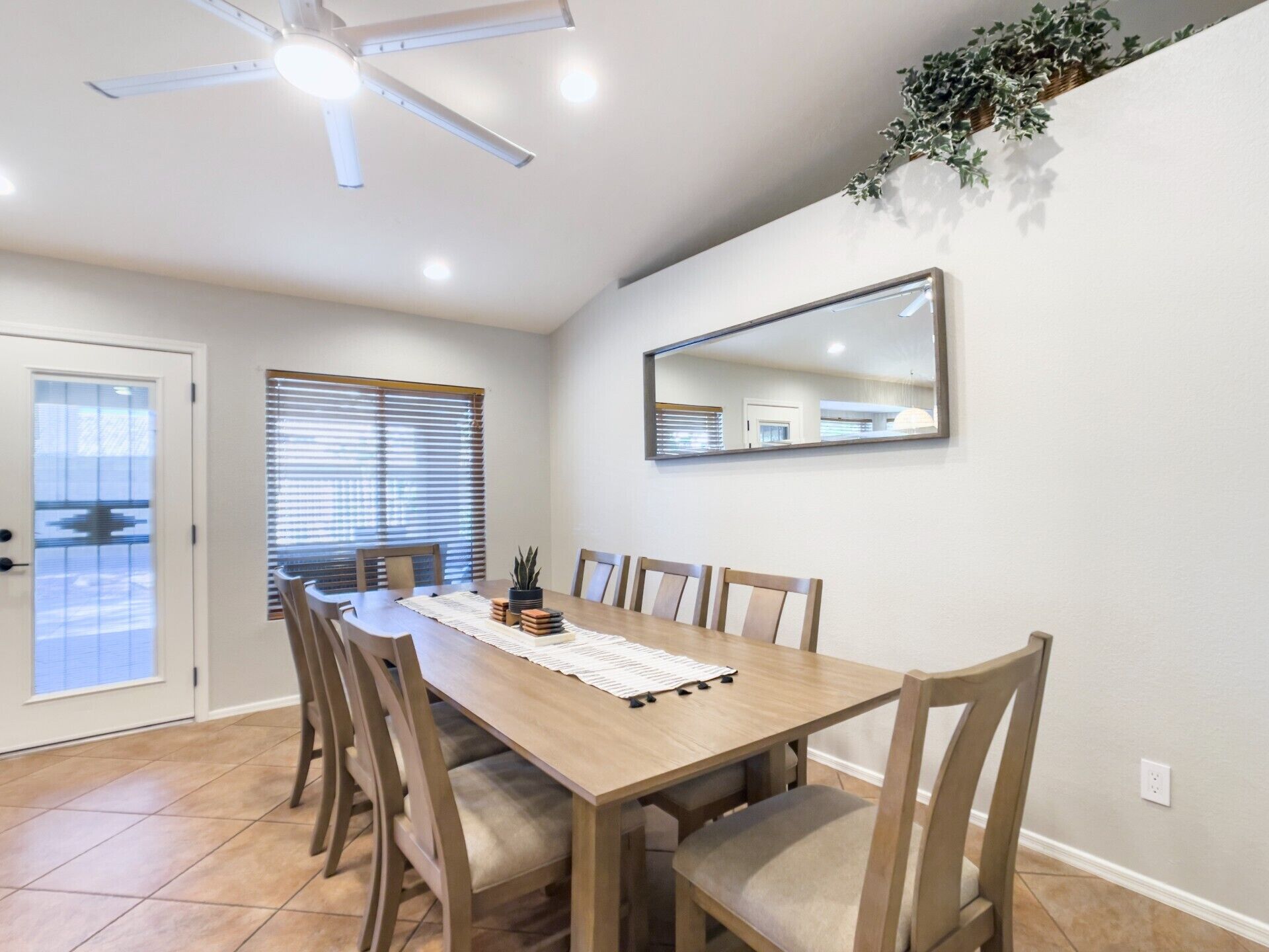 Un coin repas comprenant une table en bois, six chaises, un ventilateur de plafond, un grand miroir et une plante en pot.