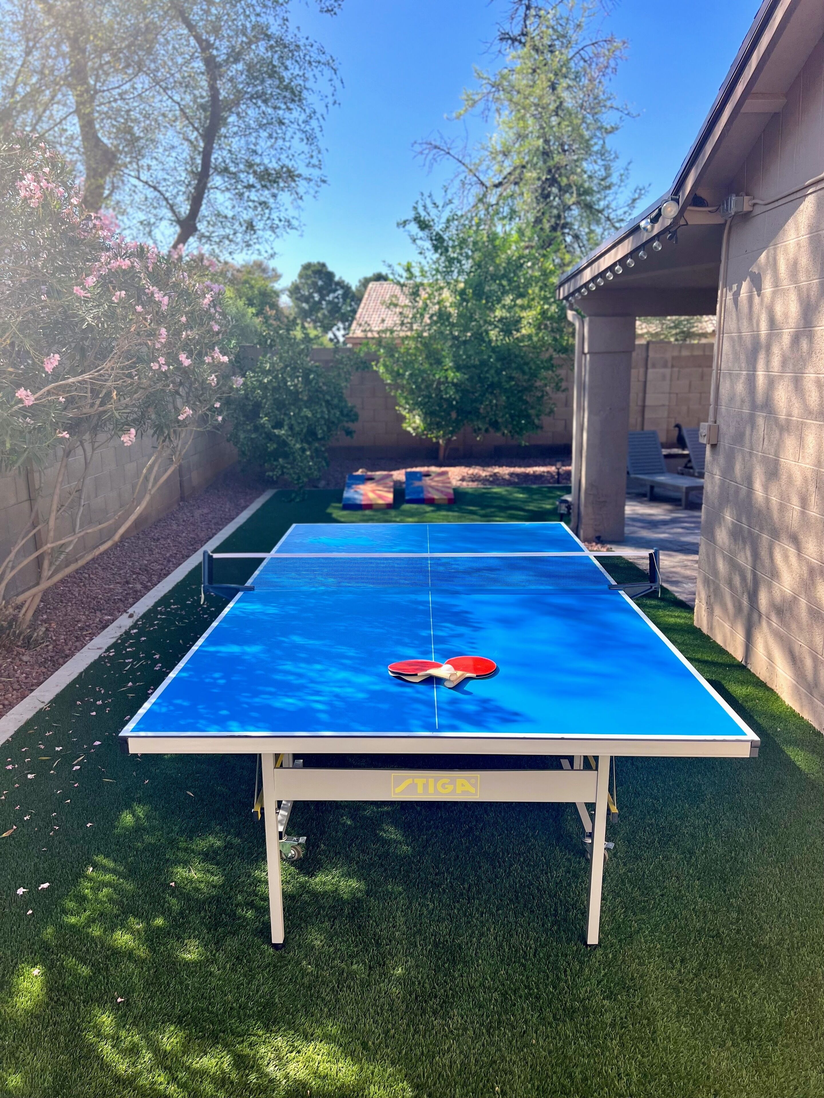 Une table de ping-pong installée dans un espace extérieur recouvert de gazon synthétique, entourée d’arbres et d’un mur en pierre.