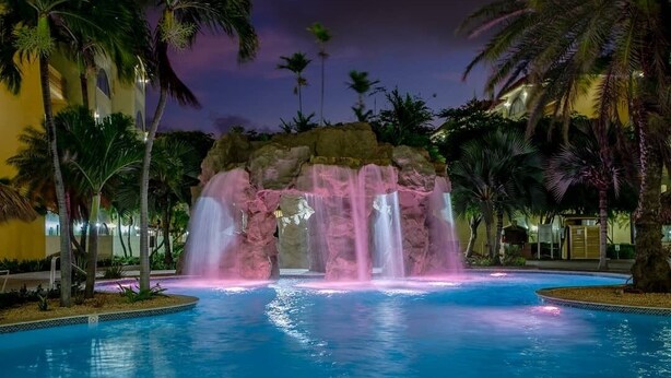 Área de piscina nocturna con una cascada central, rodeada de palmeras y un edificio al fondo.