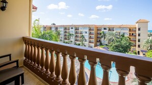 Un balcón con barandilla de madera que da a un hotel de varios pisos y balcones, piscina y palmeras.