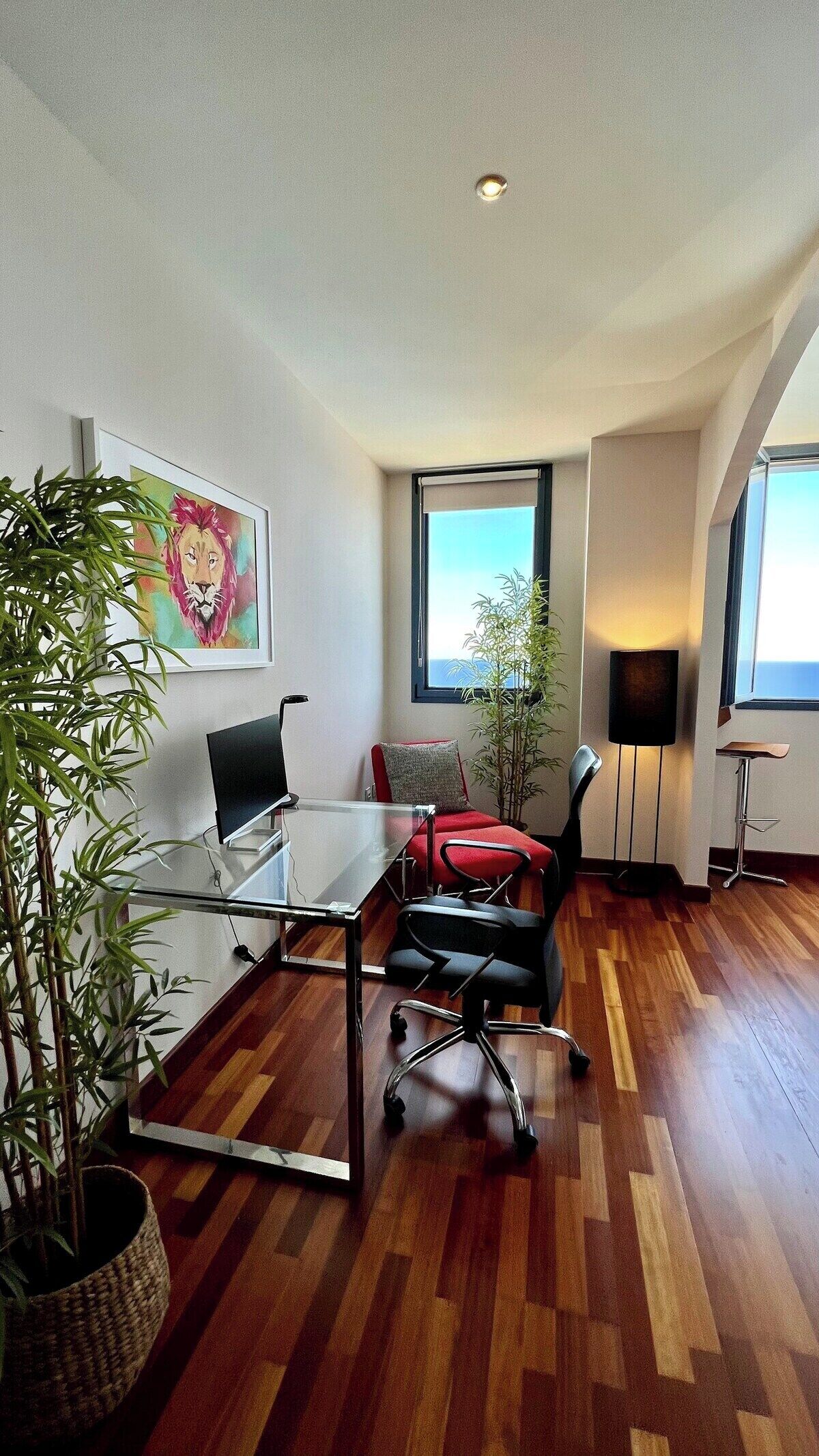 A modern office space with a glass table, black office chairs, a red cushion, a large potted plant, and a framed artwork on the wall.