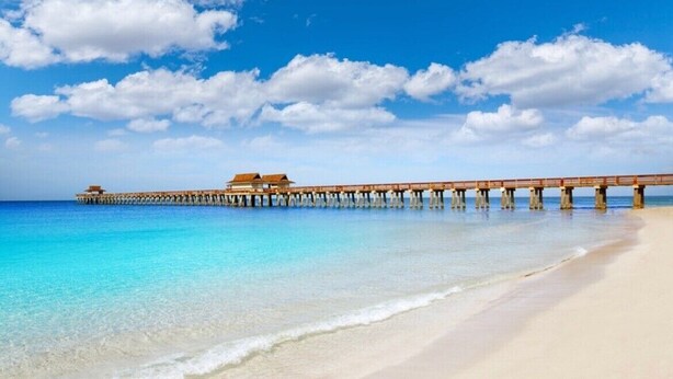 Una playa con un muelle de madera que se adentra en el océano, un pequeño edificio al final del muelle, y cielos azules despejados con algunas nubes.