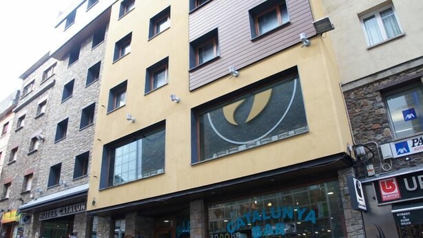 Een meerhoog gebouw met een groot raam waarop een logo, een bord met 'CATALUNYA BAR' en een 'PATRIZIA AUTOMOBILI' bord staan.