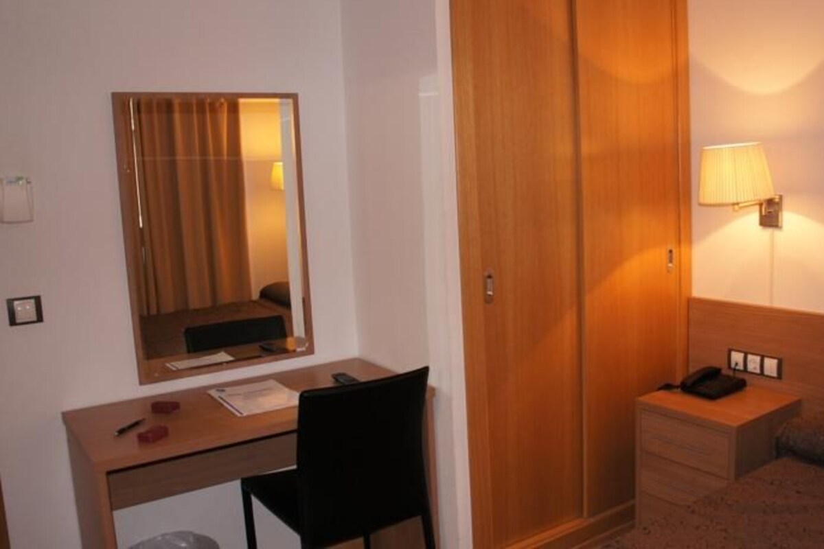 Habitación de hotel con escritorio, silla, mesita de noche con lámpara, armario y cama.