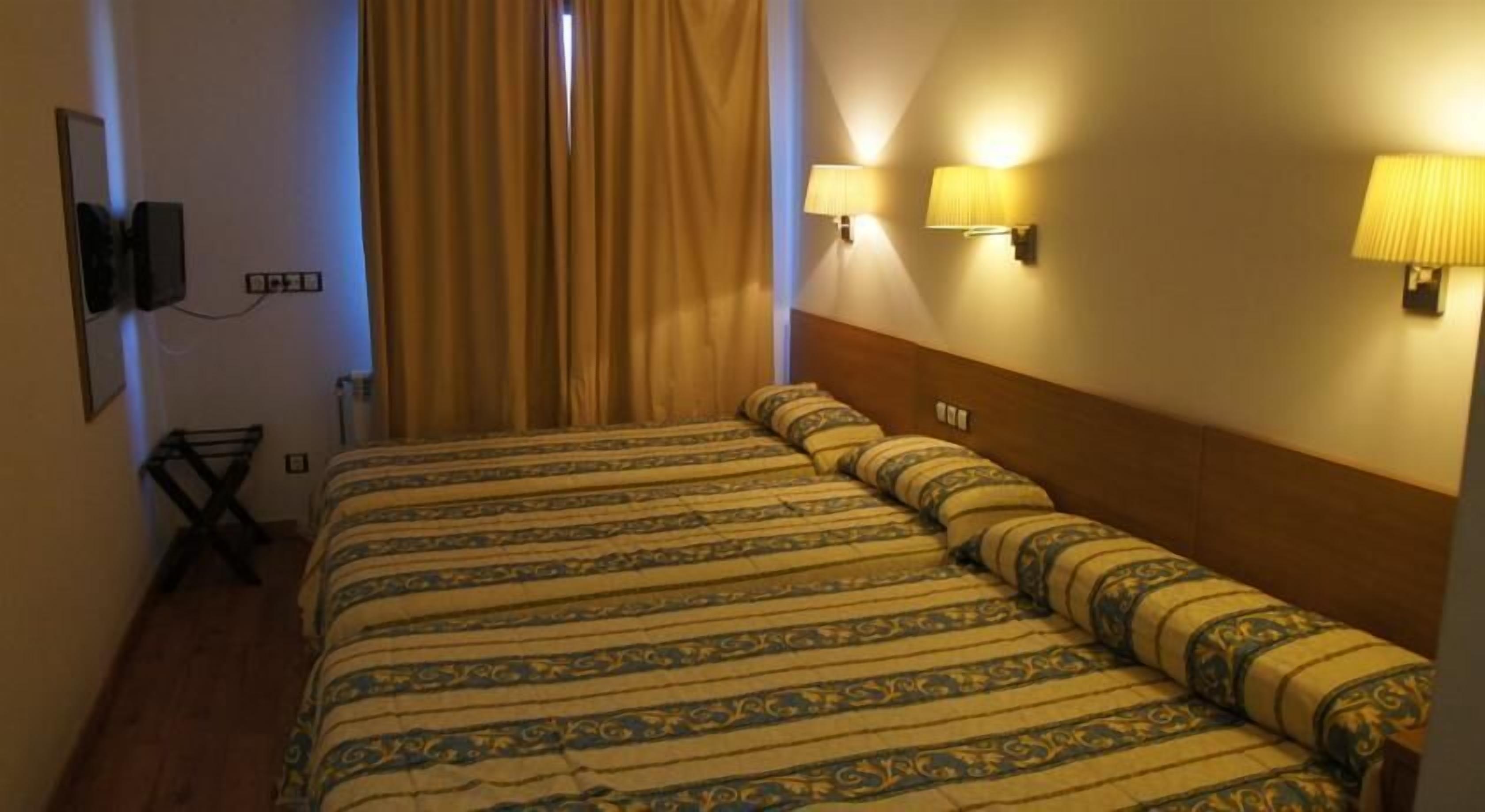 A hotel room with a large bed, two wall-mounted lamps, a TV, and a window with curtains.