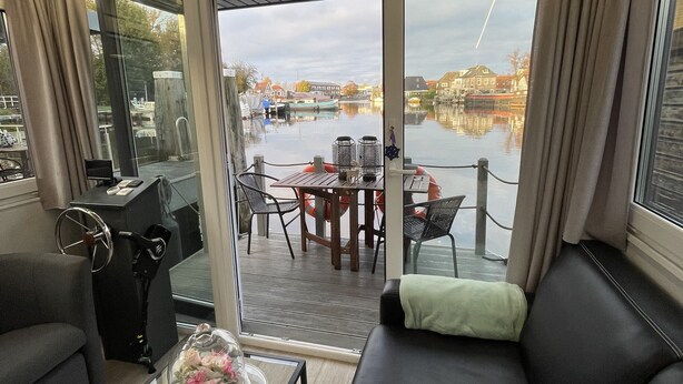 A modern living room with a sofa, a coffee table, and a view of a water body and buildings. - Sleeping on the water (Zwartsluis)