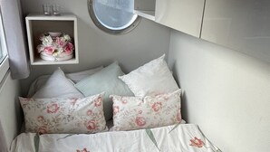 A compact bedroom with a bed, pillows, a small bedside table with a mirror, and a wall-mounted shelf with flowers. - Sleeping on the water (Zwartsluis)