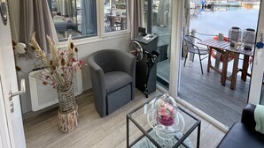 A modern interior with a grey armchair, a small table with a glass top, a wicker vase with dried plants, and a view of a dock and boats outside. - Sleeping on the water (Zwartsluis)