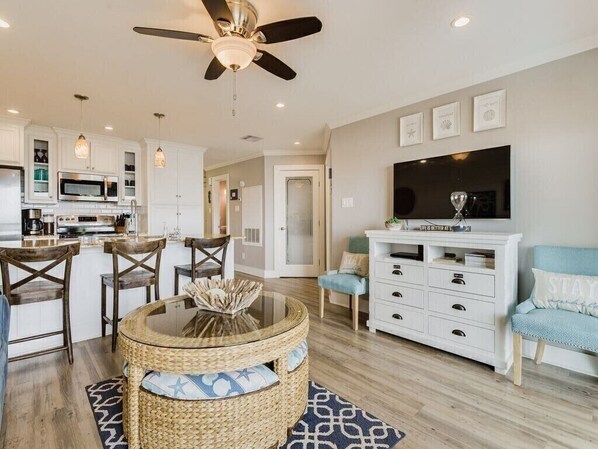 A modern living room with a round glass table, a ceiling fan, a flat-screen TV, and a white sofa.