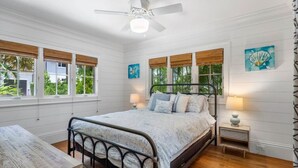 A bedroom with a bed, bedside tables, a ceiling fan, and large windows with wooden blinds. - The "Water House" Cottages (Gulf Stream)