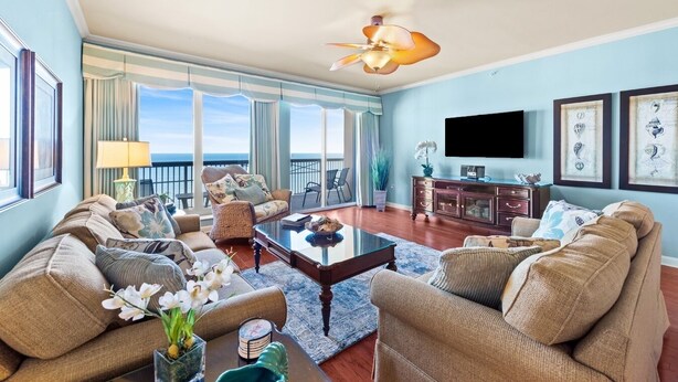 A spacious living room with a large window overlooking the ocean, a ceiling fan, and a flat-screen TV mounted on the wall. - $150 activities - E-gift card when you book - Stunning Oceanfront Retreat in Gated Community | M2303 (Myrtle Beach)