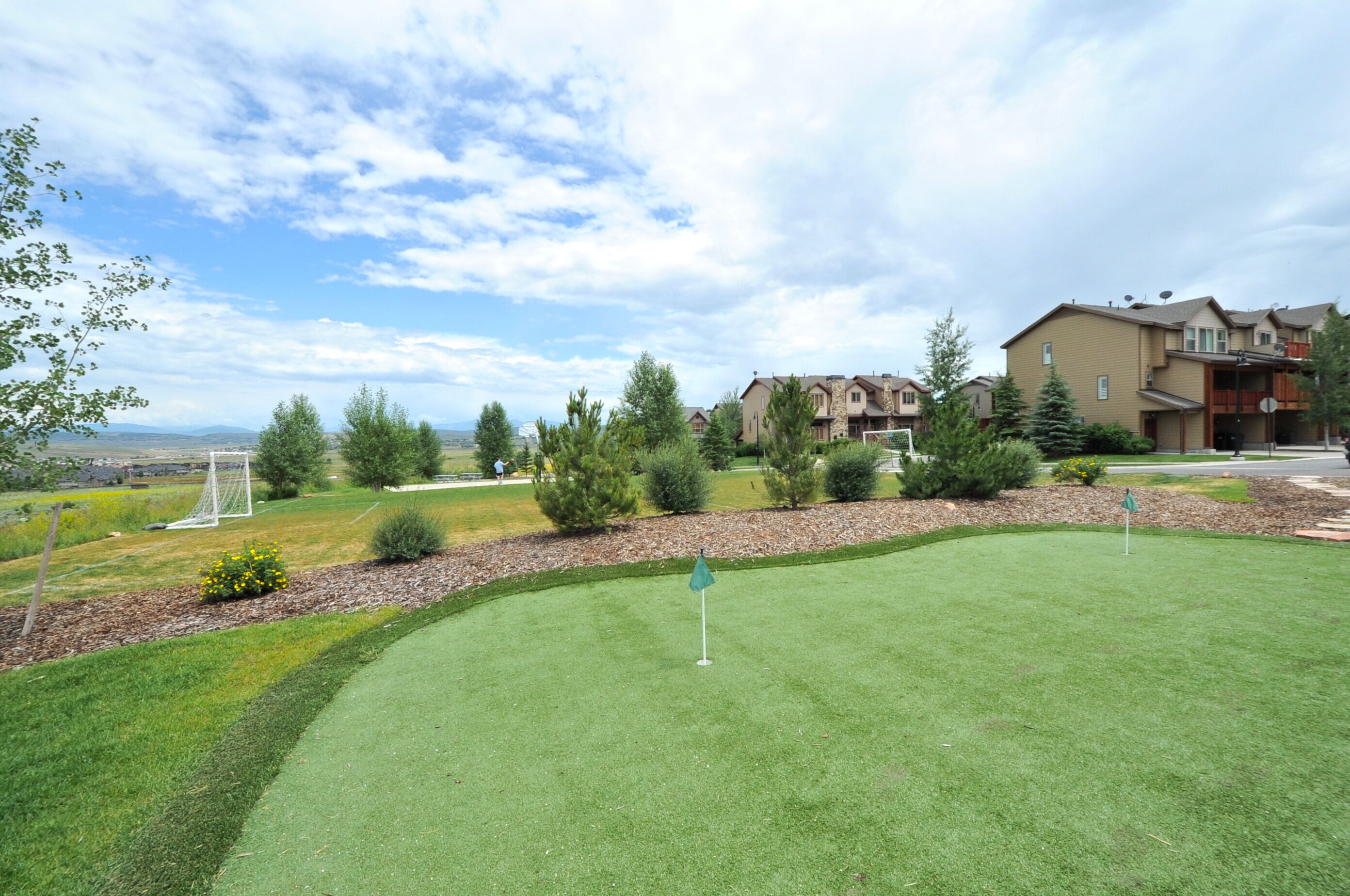 Un campo de golf bien cuidado con un putting green, rodeado de árboles maduros y una zona residencial con casas.