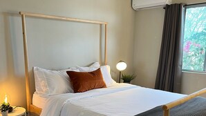 Ein ordentlich bezogenes Bett mit weißer und brauner Bettwäsche, eine Nachttischlampe und ein kleiner Tisch mit einer Pflanze.