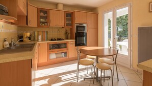A modern kitchen with wooden cabinets, a built-in oven, and a dining area with a table and chairs. - Lenerie - 800m plage - Wifi - Parking (Préfailles)