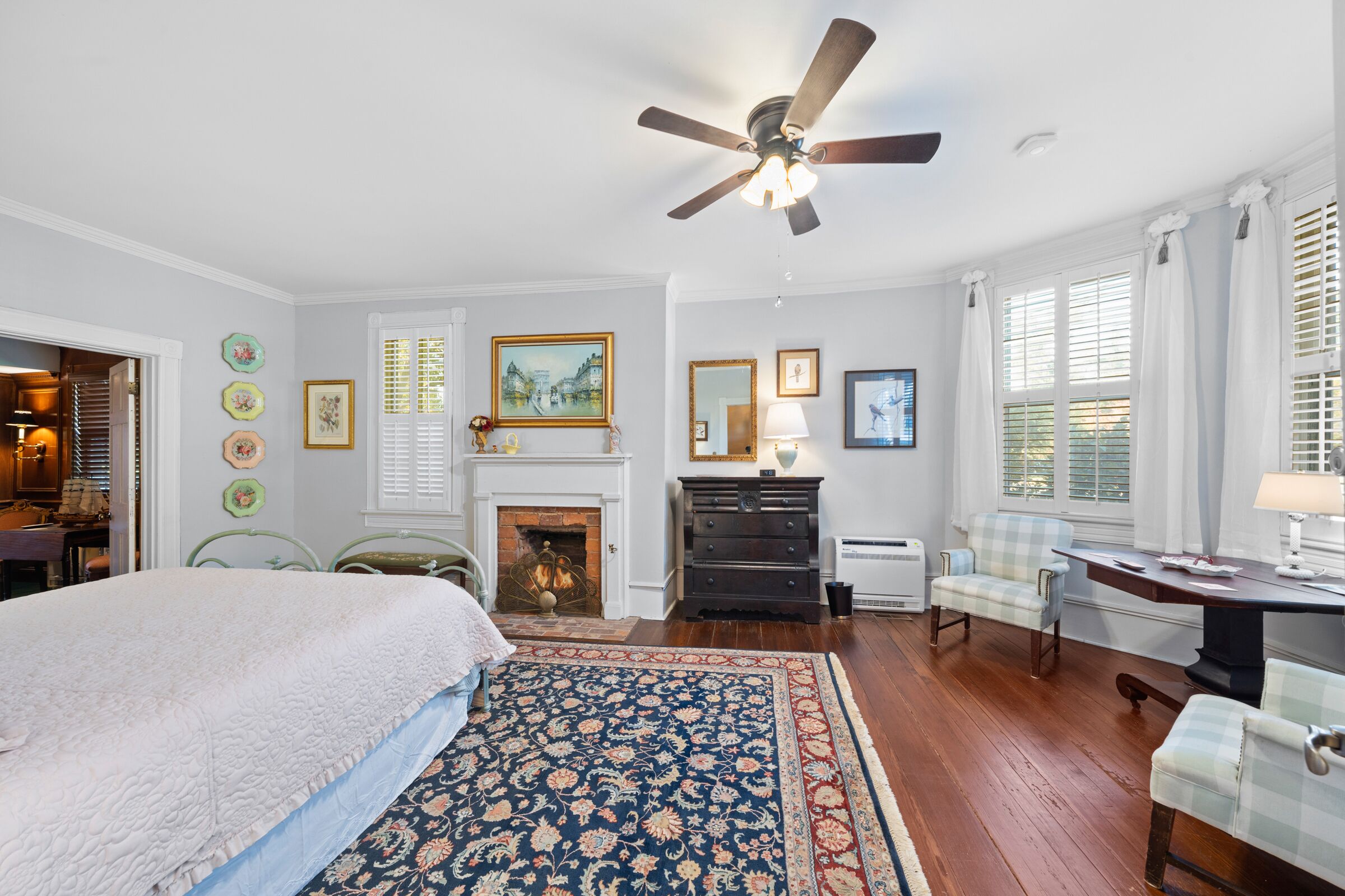 A bedroom with a large bed, a fireplace, a ceiling fan, and a view of the outside through the windows.