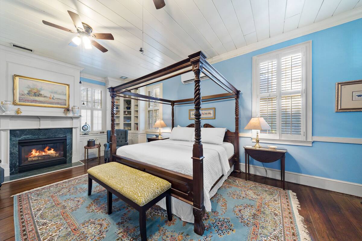A four-poster bed with a bench, a fireplace, and a large window with shutters.