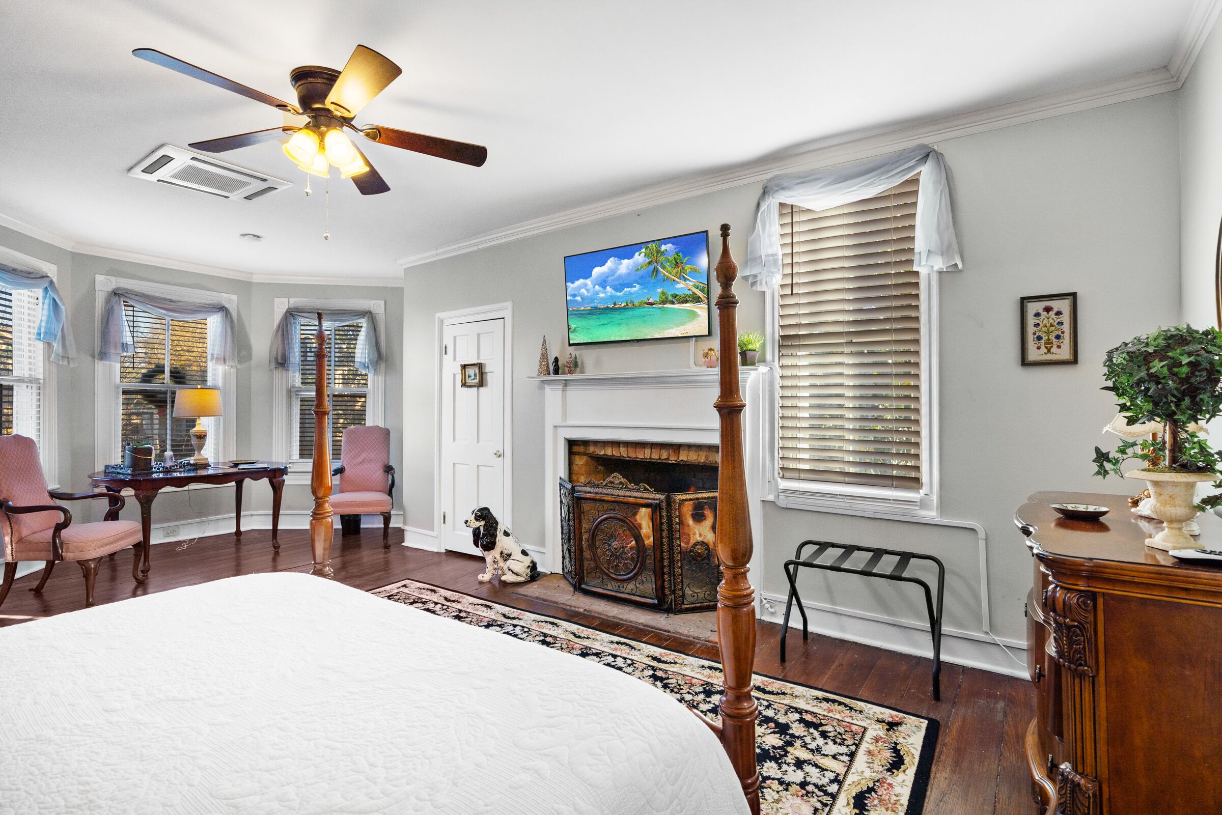 A bedroom with a large bed, a ceiling fan, a fireplace, and a TV on the wall.
