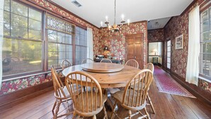 A dining room with a wooden table and chairs, large windows with floral wallpaper, and a chandelier. - The Inn on North Jefferson (Milledgeville)