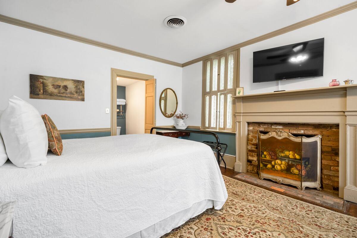 A bedroom with a large bed, a television mounted above a fireplace, and a door leading to another room.