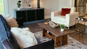 A modern living room with a dark grey sofa, a white armchair, a wooden coffee table, and a blue patterned rug. There is a black entertainment unit with decorative items and a lamp.