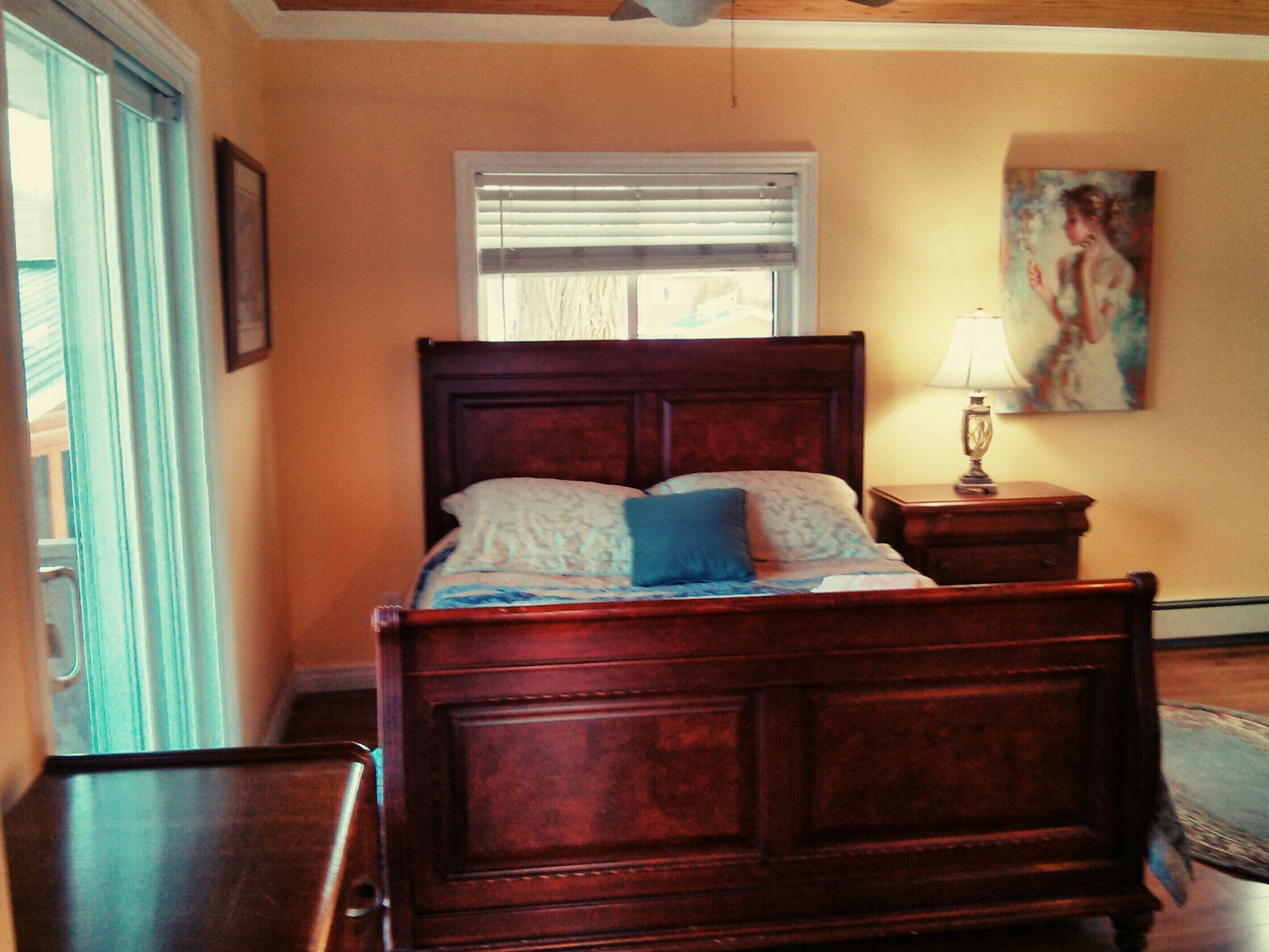 Un dormitorio con cama de madera, una ventana con persianas, una mesita de noche con lámpara y un cuadro en la pared.